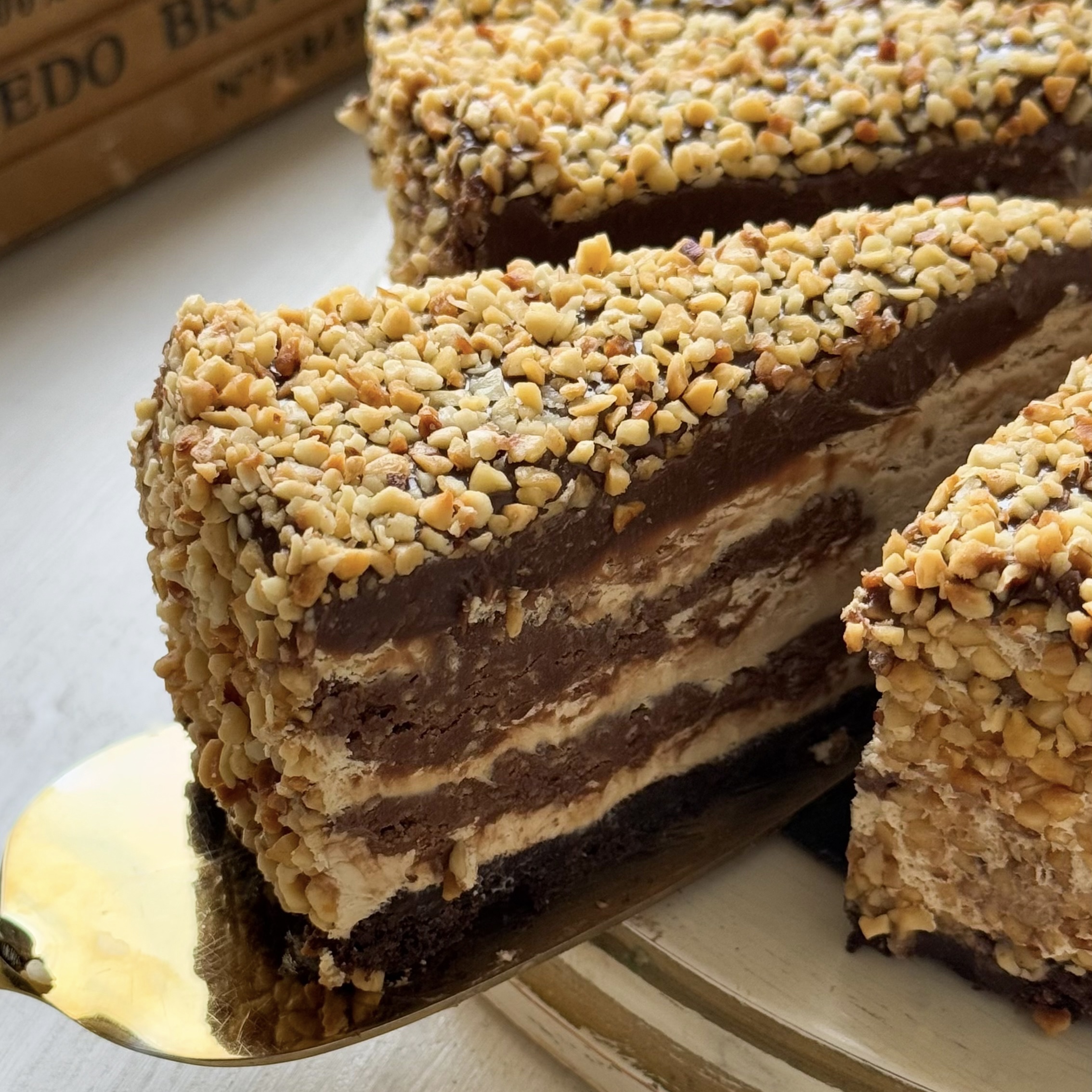 Нежный торт с сердцевиной из фундучного пралине, молочного шоколада, крема из взбитых сливок и хрустящей крошки на тонкой шоколадной подложке. Торт залит толстым шоколадным слоем и украшен дроблёным орехом. изображение 3