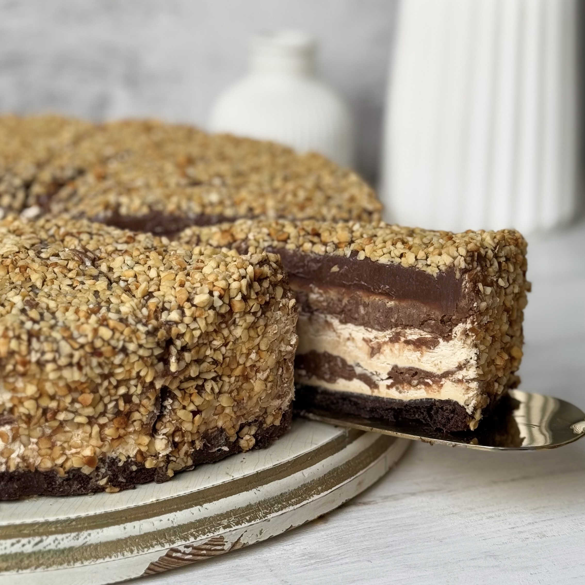 Нежный торт с сердцевиной из фундучного пралине, молочного шоколада, крема из взбитых сливок и хрустящей крошки на тонкой шоколадной подложке. Торт залит толстым шоколадным слоем и украшен дроблёным орехом. изображение 8